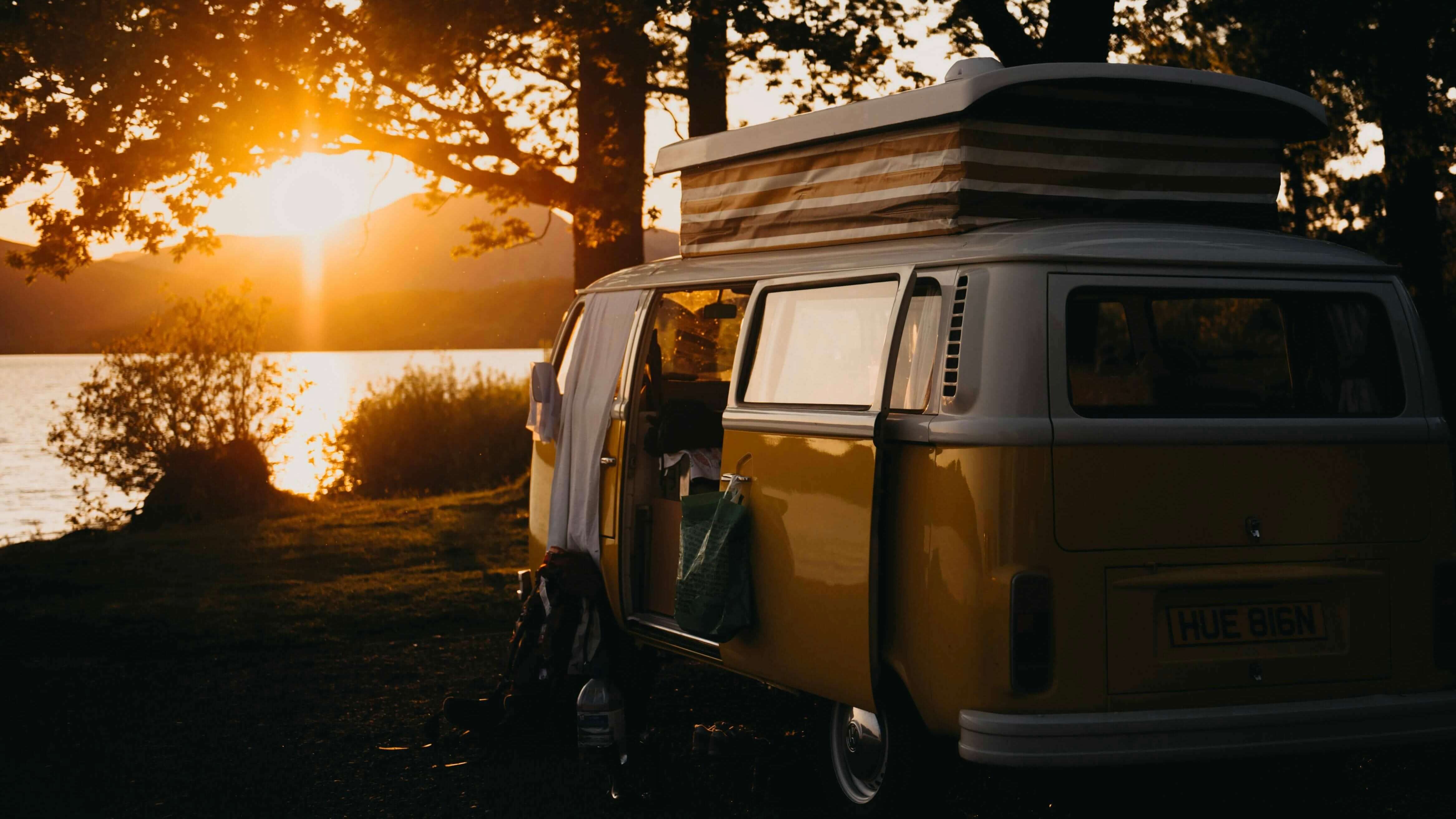 VW Bulli Campervan am Seeufer bei Sonnenuntergang mit offener Schiebetür und aufgeklapptem Dach